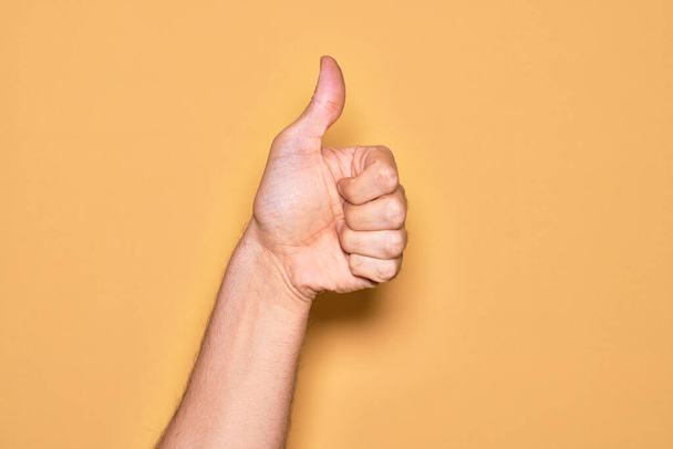 Hand of caucasian young man showing fingers over isolated yellow background doing successful approval gesture with thumbs up, validation and positive symbol - Photo, Image