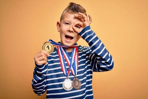 Junge kleine kaukasische Siegerkind trägt Preisverleihung Medaillen über gelbem Hintergrund mit glücklichem Gesicht lächelnd tun ok Zeichen mit Hand auf Auge Blick durch die Finger - Foto, Bild