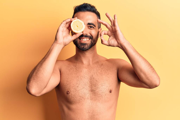 Fiatal spanyol férfi áll félmeztelen gazdaság citrom csinál rendben jel ujjaival, mosolygós barátságos gesztus kiváló szimbólum  - Fotó, kép