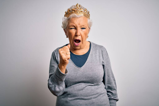 Senior beautiful grey-haired woman wearing golden queen crown over white background angry and mad raising fist frustrated and furious while shouting with anger. Rage and aggressive concept. - Photo, Image
