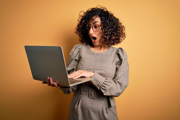 驚きの顔でショックで怖がって黄色の背景の上にノートパソコンを使用して作業眼鏡をかけて美しいアラブのビジネス女性,恐怖の表現で恐れと興奮 - 写真・画像