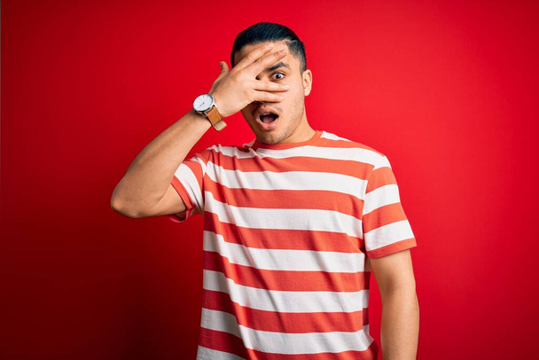 Молодой бразилец в повседневной полосатой футболке, стоящий на изолированном красном фоне, заглядывая в ударное закрывающее лицо и глаза рукой, глядя сквозь пальцы со смущенным выражением лица
. - Фото, изображение