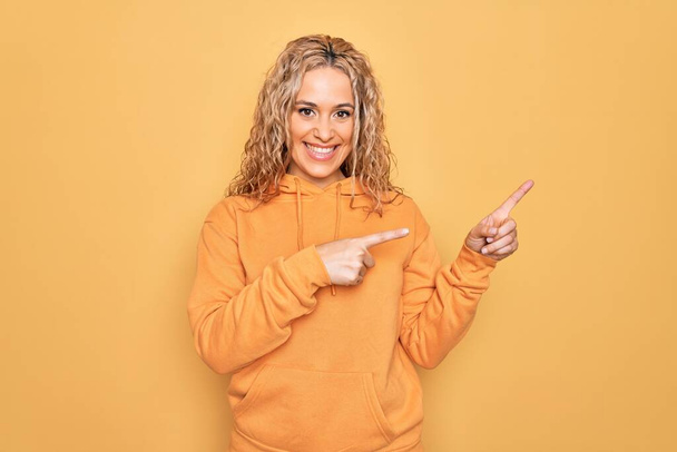 Jonge mooie blonde sportieve vrouw dragen casual sweatshirt over gele achtergrond glimlachen en kijken naar de camera wijzen met twee handen en vingers naar de zijkant. - Foto, afbeelding