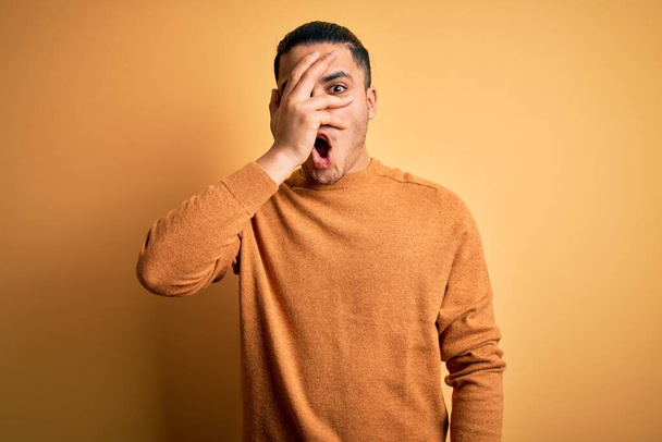 Jeune homme brésilien portant pull décontracté debout sur fond jaune isolé regardant dans le choc couvrant le visage et les yeux avec la main, regardant à travers les doigts avec une expression embarrassée. - Photo, image