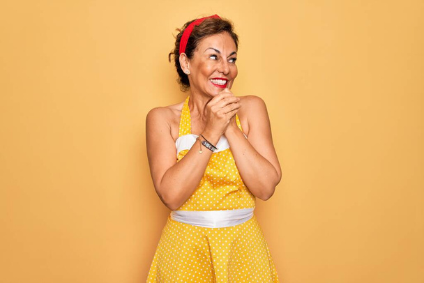 Középkorú idős pin up nő visel 50-es évek stílus retro ruha sárga háttér nevetés ideges és izgatott a kezét az állára néz oldalra - Fotó, kép