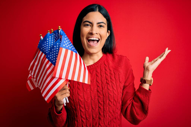 Fiatal gyönyörű spanyol nő kezében Amerikai zászló függetlenség napján nagyon boldog és izgatott, győztes kifejezés ünneplő győzelem sikoltozik nagy mosollyal és felemelt kézzel - Fotó, kép
