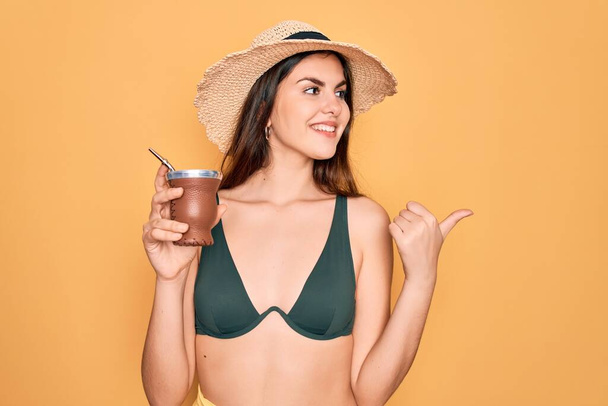 Young beautiful girl wearing swimwear bikini and summer hat drinking traditional mate drink pointing and showing with thumb up to the side with happy face smiling - Foto, imagen