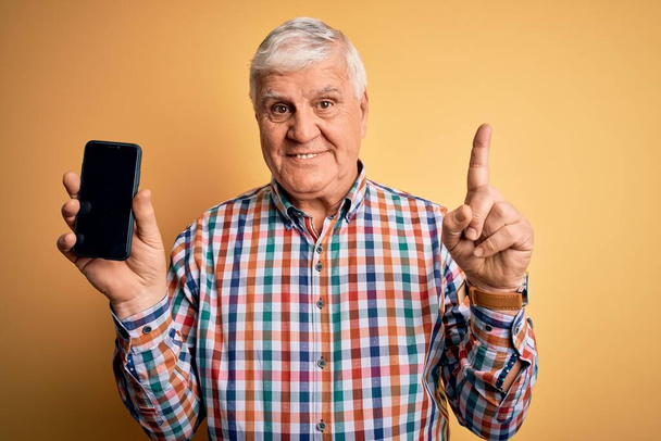 Senior bello hoary uomo che tiene smartphone mostrando schermo su sfondo giallo sorpreso con un'idea o una domanda che punta il dito con la faccia felice, numero uno - Foto, immagini