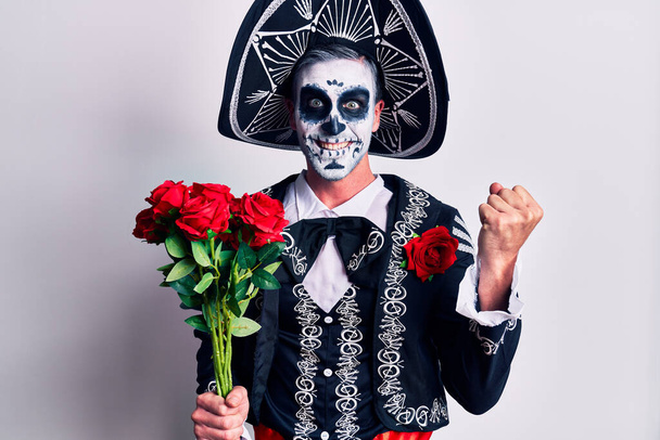 Young man wearing mexican day of the dead costume holding flowers screaming proud, celebrating victory and success very excited with raised arms  - Photo, Image