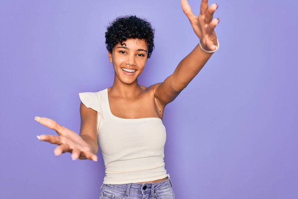 Jonge mooie Afrikaanse Amerikaanse afro vrouw draagt casual t-shirt over paarse achtergrond kijkend naar de camera glimlachend met open armen voor knuffel. Vrolijke uitdrukking die geluk omarmt. - Foto, afbeelding