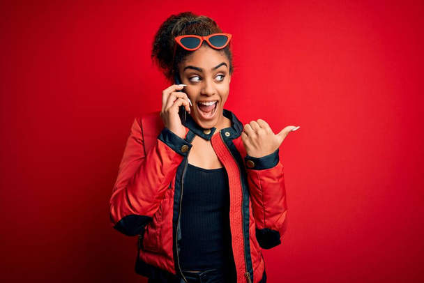 Jovem menina afro-americana ter conversa falando no smartphone sobre fundo vermelho apontando e mostrando com o polegar até o lado com o rosto feliz sorrindo
 - Foto, Imagem