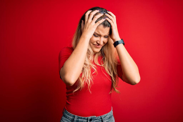 Junge schöne blonde Frau in lässigem T-Shirt, die vor isoliertem roten Hintergrund steht und unter Kopfschmerzen leidet, verzweifelt und gestresst wegen Schmerzen und Migräne. Hände auf dem Kopf. - Foto, Bild