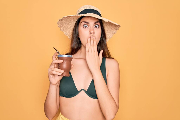 Young beautiful girl wearing swimwear bikini and summer hat drinking traditional mate drink cover mouth with hand shocked with shame for mistake, expression of fear, scared in silence, secret concept - Photo, Image