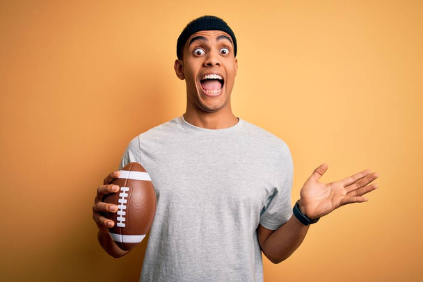若いハンサムなアフリカ系アメリカ人の男が孤立した黄色の背景の上にラグビーボールを保持非常に幸せと興奮,大きな笑顔で叫んで勝利を祝う勝者式と手を上げ - 写真・画像