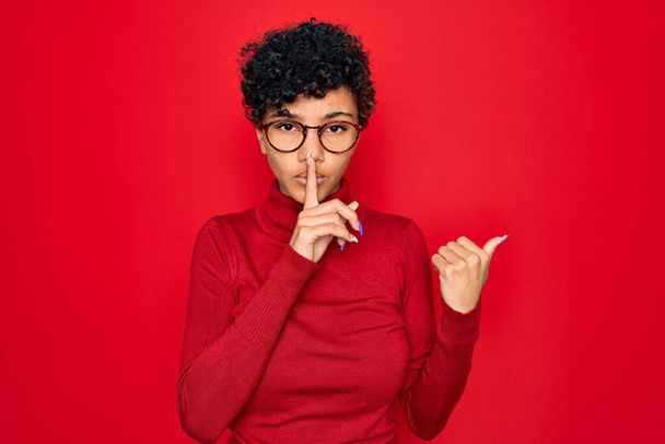Young beautiful african american afro woman wearing turtleneck sweater and glasses asking to be quiet with finger on lips pointing with hand to the side. Silence and secret concept. - Photo, Image