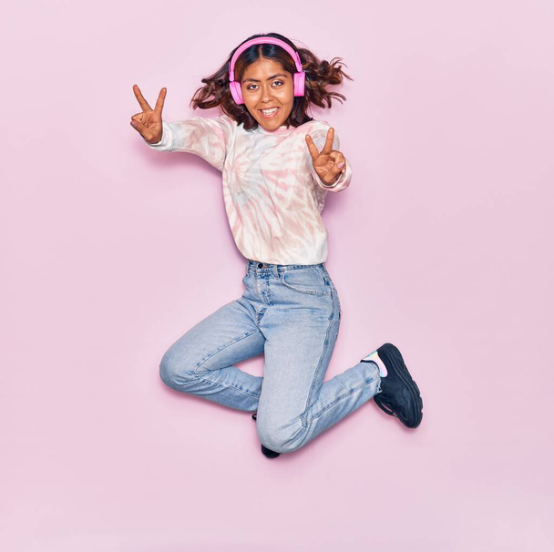 Молода красива латинка слухає музику, використовуючи навушники, усміхаючись щасливою. Стрибки з посмішкою на обличчі роблять знак перемоги над ізольованим рожевим тлом
 - Фото, зображення