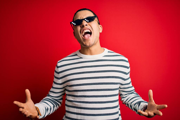 Mladý brazilský muž v legračních gorilských slunečních brýlích na izolovaném červeném pozadí, šílený a šílený, křičící a křičící s agresivním výrazem a zdviženýma rukama. Koncept frustrace. - Fotografie, Obrázek