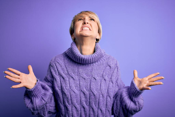 Mujer rubia joven con el pelo corto con suéter de cuello alto de invierno sobre fondo púrpura loco y loco gritando y gritando con expresión agresiva y los brazos levantados. Concepto de frustración
. - Foto, Imagen