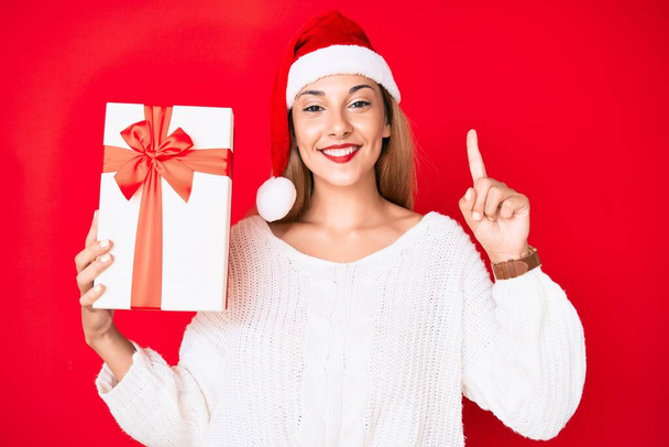 Молодая брюнетка в рождественской шляпе и с подарком в руках удивлена идеей или вопросом, указывая пальцем со счастливым лицом, номер один  - Фото, изображение
