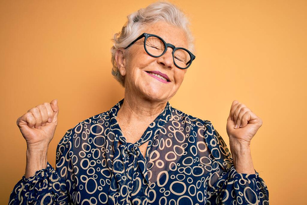 Starsza piękna siwowłosa kobieta w luźnej koszuli i okularach na żółtym tle bardzo szczęśliwa i podekscytowana wykonując gest zwycięzcy z podniesionymi ramionami, uśmiechnięta i krzycząca o sukces. Koncepcja uroczystości. - Zdjęcie, obraz