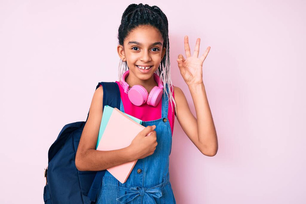 Νεαρό αφροαμερικανό κορίτσι παιδί με κοτσίδες κρατώντας σακίδιο μαθητή και βιβλία κάνει ok υπογράψει με τα δάχτυλα, χαμογελώντας φιλικό gesturing εξαιρετικό σύμβολο  - Φωτογραφία, εικόνα