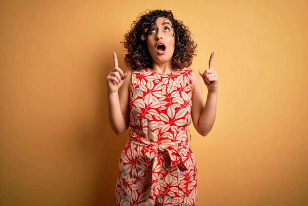 Young beautiful curly arab woman on vacation wearing summer floral dress and sunglasses amazed and surprised looking up and pointing with fingers and raised arms. - Photo, Image