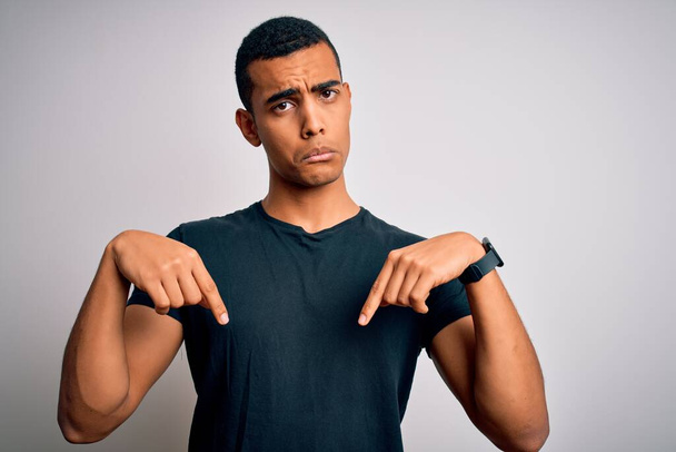 Junger gutaussehender afrikanisch-amerikanischer Mann in lässigem T-Shirt vor weißem Hintergrund Zeigt traurig und aufgebracht nach unten, zeigt mit den Fingern Richtung an, unglücklich und deprimiert. - Foto, Bild