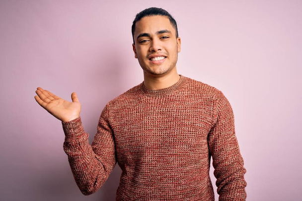 Mladý brazilský muž na sobě ležérní svetr stojící nad izolované růžové pozadí s úsměvem veselý prezentaci a ukazuje dlaní ruky při pohledu na fotoaparát. - Fotografie, Obrázek