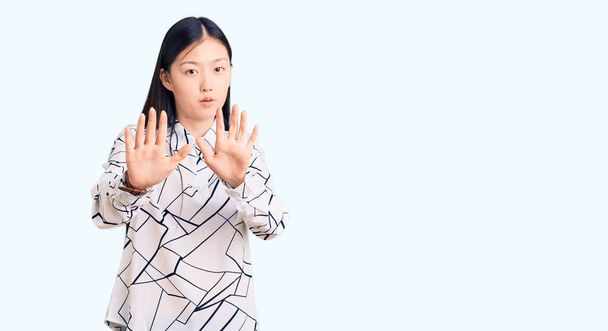 Młoda piękna Chinka ubrana w luźną koszulę odsuwa dłonie pokazując odmowę i zaprzeczenie ze strasznym i obrzydliwym wyrazem twarzy. zatrzymać i zabronione.  - Zdjęcie, obraz