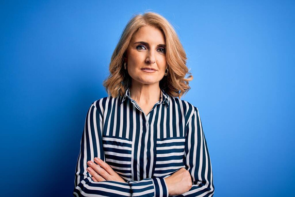 Middle age beautiful blonde woman wearing casual striped shirt standing over blue background happy face smiling with crossed arms looking at the camera. Positive person. - Fotografie, Obrázek