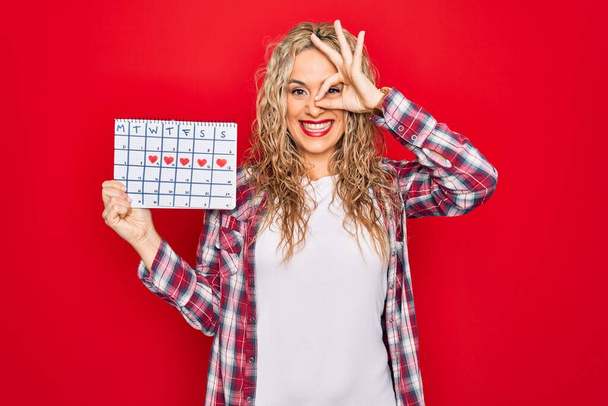 Молодая красивая блондинка держит календарь период контроля менструального цикла улыбаясь счастливо делает хорошо знак с рукой на глазу глядя сквозь пальцы
 - Фото, изображение