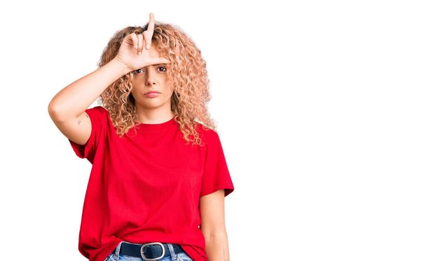 jonge blonde vrouw met krullend haar dragen casual rood tshirt maken plezier van mensen met vingers op het voorhoofd doen verliezer gebaar spotten en beledigend.  - Foto, afbeelding