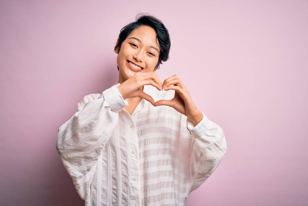 Junge schöne asiatische Mädchen in lässigem Hemd, das über isoliertem rosa Hintergrund steht und verliebt lächelnd Herzsymbolform mit Händen macht. Romantisches Konzept. - Foto, Bild