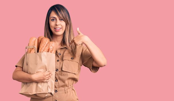 Молодая красивая женщина держит бумажный пакет с хлебом указывая пальцем на один себя улыбается счастливый и гордый  - Фото, изображение