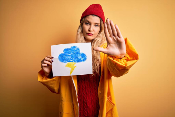 Young beautiful blonde woman wearing raincoat holding banner with cloud thunder image with open hand doing stop sign with serious and confident expression, defense gesture - Фото, зображення