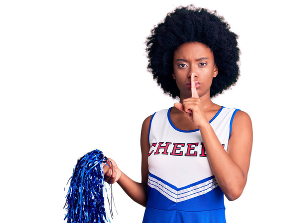 Jeune femme afro-américaine portant un uniforme de pom-pom girl tenant un pompon demandant de se taire avec le doigt sur les lèvres. silence et concept secret.  - Photo, image