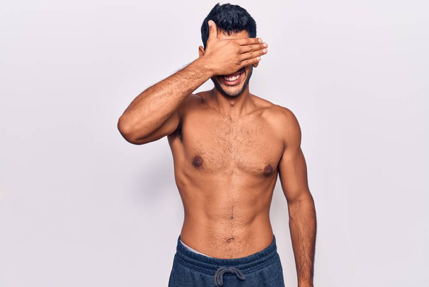 Young latin man standing shirtless smiling and laughing with hand on face covering eyes for surprise. blind concept.  - Foto, Imagem