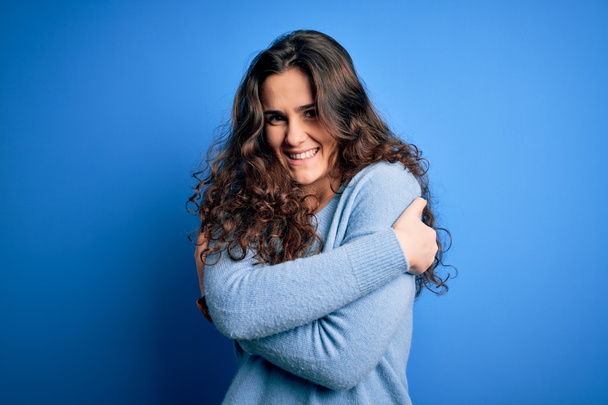 Young beautiful woman with curly hair wearing blue casual sweater over isolated background Hugging oneself happy and positive, smiling confident. Self love and self care - Photo, Image