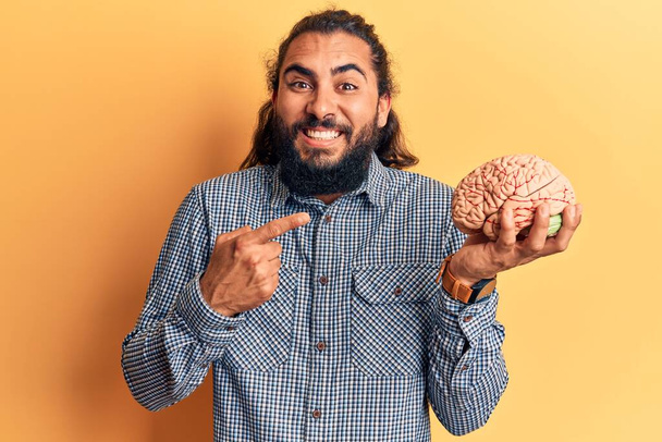 Jonge Arabier man houdt de hersenen glimlachend vrolijk wijzend met hand en vinger  - Foto, afbeelding