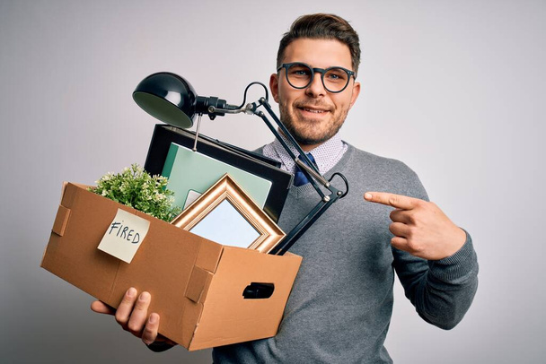 Joven hombre de negocios con ojos azules sosteniendo la caja de la oficina siendo despedido del trabajo muy feliz señalando con la mano y el dedo
 - Foto, Imagen