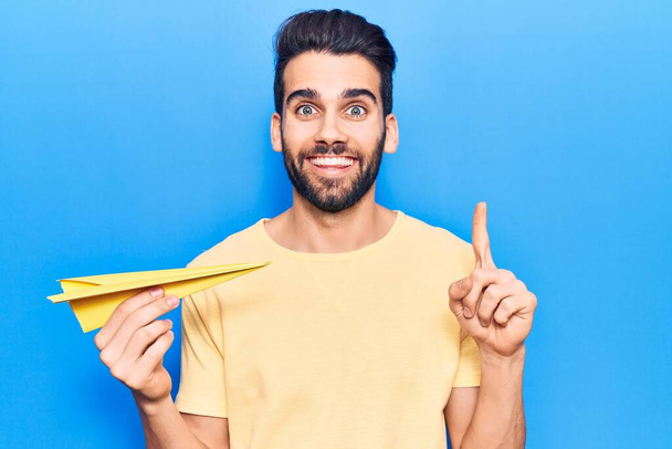 Young handsome man with beard holding yellow paper airplane smiling with an idea or question pointing finger with happy face, number one  - Fotó, kép