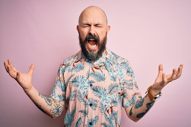 Hombre calvo guapo con barba y tatuaje con camisa floral casual sobre fondo rosa loco y loco gritando y gritando con expresión agresiva y los brazos levantados. Concepto de frustración
. - Foto, imagen