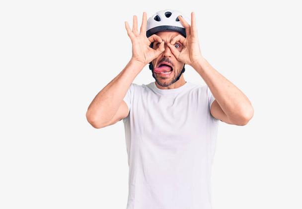 自転車のヘルメットをかぶっている若いハンサムな男は、双眼鏡が舌を突き出しているようなokのジェスチャーをして、目は指を見ている。クレイジーな表現.  - 写真・画像