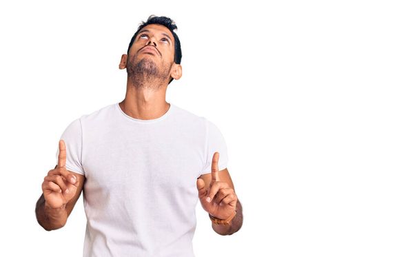 Jeune homme latino portant des vêtements décontractés pointant vers le haut l'air triste et contrarié, indiquant la direction avec les doigts, malheureux et déprimé.  - Photo, image