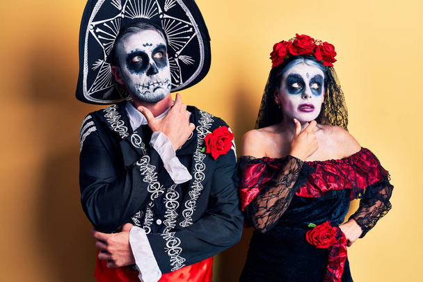 Young couple wearing mexican day of the dead costume over yellow thinking worried about a question, concerned and nervous with hand on chin  - Photo, Image