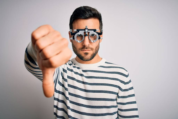 Jonge knappe man met baard draagt optometrie bril over geïsoleerde witte achtergrond kijken ongelukkig en boos tonen afwijzing en negatief met duimen naar beneden gebaar. Slechte uitdrukking. - Foto, afbeelding