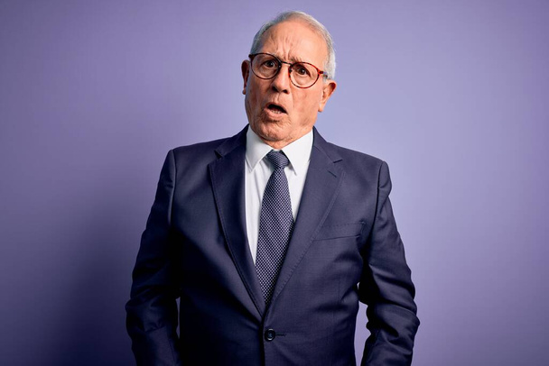 Grey haired senior business man wearing glasses and elegant suit and tie over purple background In shock face, looking skeptical and sarcastic, surprised with open mouth - Photo, Image
