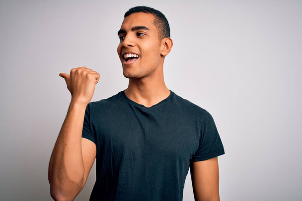 Fiatal jóképű afro-amerikai férfi alkalmi pólóban áll a fehér háttér felett mosolyogva boldog arccal, és mutatva oldalra hüvelykujjával felfelé. - Fotó, kép