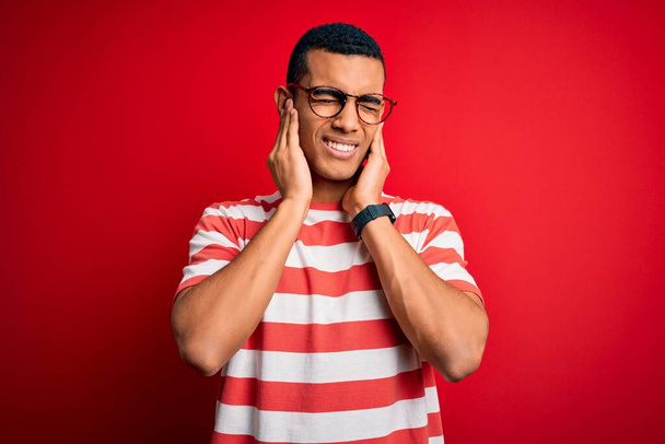 カジュアルなストライプのTシャツを着た若いハンサムなアフリカ系アメリカ人男性と、大きな音楽のノイズのためにいらいらする表情で指で耳を覆う眼鏡。ろう者の概念. - 写真・画像