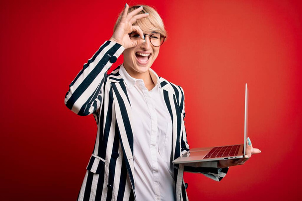 Jeune femme d'affaires blonde avec les cheveux courts portant des lunettes de travail à l'aide d'un ordinateur portable avec le visage heureux souriant faire ok signe avec la main sur les yeux regardant à travers les doigts - Photo, image